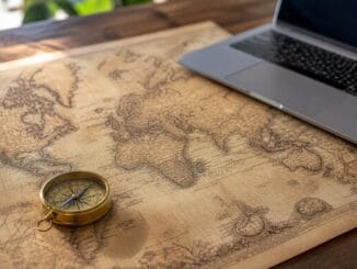 Antique world map with a brass compass on a wooden desk beside a laptop